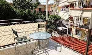 Balkong med glassbord og to metallstoler, med utsikt over grøntområder og omkringliggende bygninger. (AI-generert tekst)