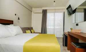 Et hotellrom med en dobbeltseng med hvitt sengetøy og gult sengeteppe, en sofa med puter, et skrivebord med speil og en TV på veggen. Balkongdører med lyse gardiner. (AI-generert tekst)