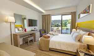 Et hotellrom med seng, skrivebord, TV og en balkong som viser en grønn utsikt. (AI-generert tekst)