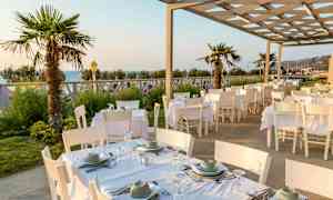 Uteservering med hvite bord og stoler, palmetrær og havutsikt. (AI-generert tekst)