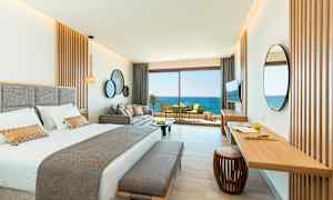 Et stilrent hotellrom med en dobbeltseng, sofa og et skrivebord. Store glassdører åpner seg mot en balkong med utsikt over havet. (AI-generert tekst)