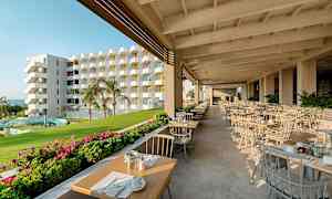 En utendørs restaurant med dekkede bord, grøntområder og en flerfarget bygning i bakgrunnen. (AI-generert tekst)