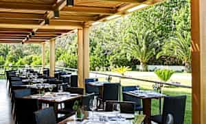 En utendørs restaurant med dekkede bord og utsikt over en hage med palmer. (AI-generert tekst)