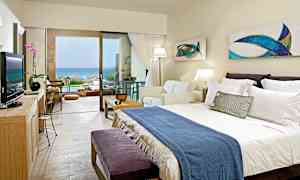 Et hotellrom med dobbeltseng, sofa og stor glassdør som fører til en terrasse med utsikt over havet. (AI-generert tekst)