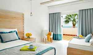 Et lyst og luftig hotellrom med seng, trepaneler, blomster på et sidebord og åpne glassdører som fører til en balkong med havutsikt. (AI-generert tekst)