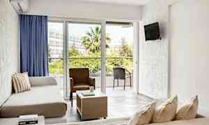 Et lyst hotellrom med sittegruppe, sofaer, en stol, et salongbord og store vinduer som vender ut mot en balkong med utemøbler i et frodig miljø. (AI-generert tekst)