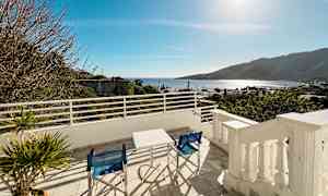 Utsikt fra en terrasse med bord og stoler, med hav og fjell i bakgrunnen. (AI-generert tekst)