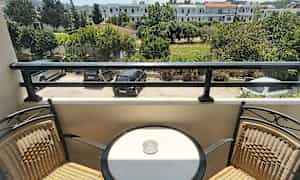 En balkong med to stoler og et rundt bord med en askebeger, med utsikt over en parkeringsplass og grønne omgivelser. (AI-generert tekst)