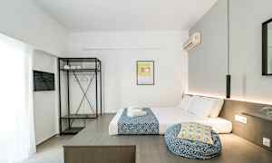 Et moderne hotellrom med en seng oppredd med hvite og blå tekstiler, en åpen garderobe med hyller og kleshengere samt en veggmontert klimaanlegg. (AI-generert tekst)