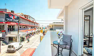 En balkong med bord, stol og blomster i forgrunnen, samt utsikt over en gate med butikker og bygninger. (AI-generert tekst)