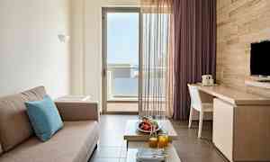 Et rom med en beige sofa, et bord med frukt og juice samt en balkongdør med utsikt. (AI-generert tekst)