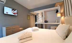 Bildet viser et hotellrom med en seng, håndklær, en TV på veggen og fasiliteter som garderobeskap, safe og vannkoker. (AI-generert tekst)