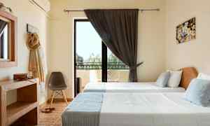 Et rom med to enkeltsenger, et stort vindu med mørke gardiner og balkongmøbler utenfor. (AI-generert tekst)