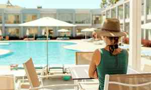En person sitter ved et bord nær et basseng med parasoller. I bakgrunnen sees en hotellbygning. (AI-generert tekst)