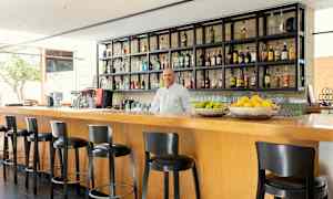 En moderne bar med høye stoler ved en trebardisk, hyller fylt med flasker og fruktkurver med sitrusfrukter. (AI-generert tekst)