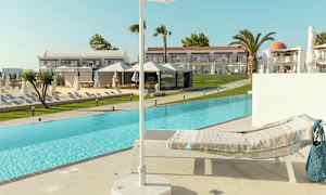 En solseng med et teppe ved kanten av et basseng, med en parasoll i forgrunnen og hotellbygninger i bakgrunnen. (AI-generert tekst)