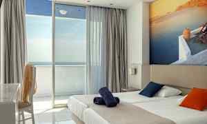 Hotellrom med dobbeltseng, fargerike puter og balkong med havutsikt. (AI-generert tekst)