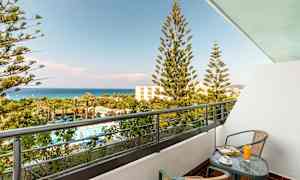 En balkong med et bord og to stoler. Utsikt over et basseng og frodig område med palmer samt havet i bakgrunnen. (AI-generert tekst)