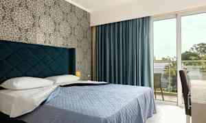 Et hotellrom med en seng, blåtteppe og blå gardiner, samt et vindu som fører til en balkong. (AI-generert tekst)