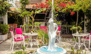 En fontene med en hvit statue av en kvinne i en frodig hage med bord og stoler. (AI-generert tekst)