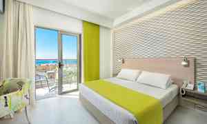 Et hotellrom med en seng, grønt sengeteppe, lyse møbler og en balkong med havutsikt. (AI-generert tekst)