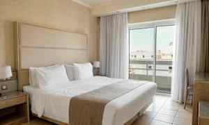 Et hotellrom med en dobbeltseng kledd i hvitt sengetøy, nattbord og store vinduer med gardiner som slipper inn lys. (AI-generert tekst)