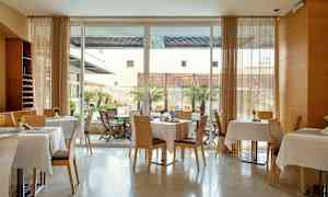 Et moderne rom med dekkede bord og stoler, samt store vinduer mot en uteplass med trær og møbler. (AI-generert tekst)