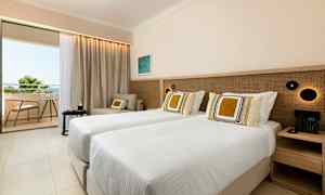 Et hotellrom med to enkeltsenger, en sittegruppe og en balkong som viser utsikt over grøntområder og himmel. (AI-generert tekst)