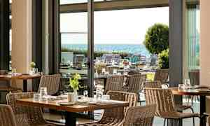 Utsikt over en moderne restaurant med flettestoler og bord, med en hage og havet i bakgrunnen. (AI-generert tekst)