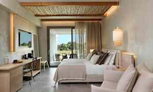 Et moderne hotellrom med en dobbeltseng, en sittegruppe og et skrivebord. Tyg slipper inn dagslys fra balkongdøren. (AI-generert tekst)