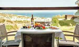 Et bord med duk, glass og en vinflaske, omgitt av stoler, med utsikt over et landskap og havet. (AI-generert tekst)