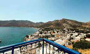 Utsikt fra en balkong med blått rekkverk over en kystby med hvite bygninger og omkringliggende fjell. (AI-generert tekst)