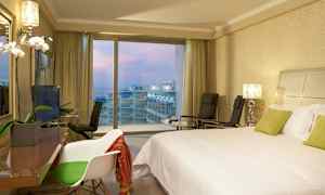 Hotellrom med en dobbeltseng, stoler og utsikt over balkonger og havet gjennom store vinduer. (AI-generert tekst)