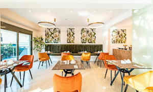 En spisesal med oransje og gule stoler samt grønn polstret benk. Bordene er dekket og dekorert. Veggene har bilder av planter. (AI-generert tekst)