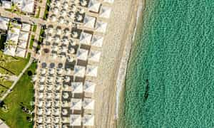 Flyfoto av en strand med solparasoller i rekker og klart turkis vann. (AI-generert tekst)