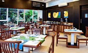 En restaurant med trebord og stoler, fargerike bordduk dekorasjoner. Bakgrunnen viser en buffet med serveringsbeholdere. Vinduer gir utsikt mot grønne planter. (AI-generert tekst)