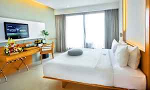 Et moderne hotellrom med en stor komfortabel seng, skrivebord med blomsteroppsats og en TV på veggen. Vinduer med lyse gardiner slipper inn dagslys. (AI-generert tekst)