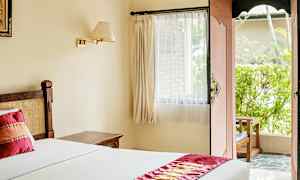 Et lyst hotellrom med en seng dekket av hvit sengetøy og en dekorativ rød løper. Et vindu med gardiner og en åpen dør fører til en uteplass med grøntområde. (AI-generert tekst)