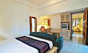 Et hotellrom med en stor seng, en liten sittegruppe, et skrivebord med speil og en gammel TV. Badet er til venstre, og det er forbindelse til et annet rom i bakgrunnen. (AI-generert tekst)