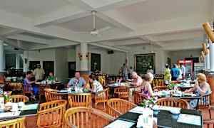 Et restaurantområde med bord og stoler, hvor folk sitter og spiser. Innredningen er lys med hvite vegger og tak. (AI-generert tekst)