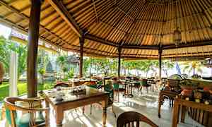 En utendørs restaurant med trebord og stoler under et halmtak, omgitt av frodig grønne områder og plassert nær stranden. (AI-generert tekst)