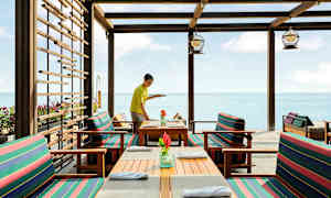 Restaurant ved sjøen med trebord og stoler med stripete puter. (AI-generert tekst)