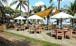 En uteservering nær en strand med trebord, stoler, parasoller og palmer. (AI-generert tekst)