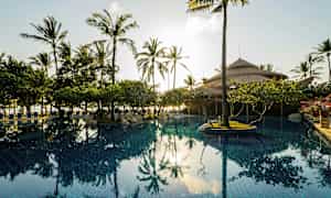 Et svømmebasseng omgitt av palmer og vegetasjon med en hytte med stråtak i bakgrunnen. (AI-generert tekst)