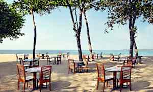 Utendørs bord og trestoler plassert på en sandstrand med havet i bakgrunnen under trær. (AI-generert tekst)