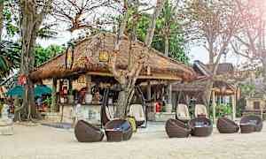 En strandrestaurant med halmtak, omgitt av trær og sitteplasser i flettede rottingmøbler. (AI-generert tekst)