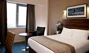 Hotellrom med seng, stol, bord og vindu med gardiner. (AI-generert tekst)
