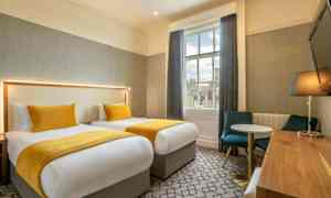 To senger med gule sengetepper og puter i et rom med bord, stoler og et vindu. (AI-generert tekst)