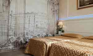 Et hotellrom med to senger dekket med gullfargede sengetepper. Bakgrunnsveggen har et kunstverk med arkitektoniske søyler og bygninger. En nattlampe og en plante står på nattbordet. (AI-generert tekst)