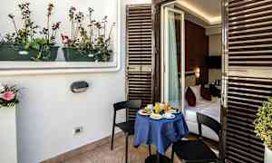 En balkong med et lite bord dekket for frokost og to stoler. Bordet er dekket med en blå duk og det leder inn til et soverom. (AI-generert tekst)